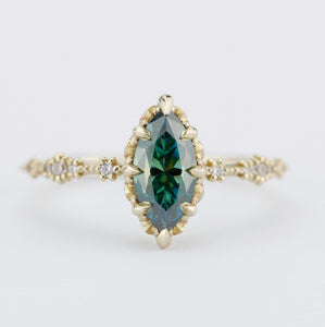 An 18k gold ring featuring a marquise-cut 10x5mm teal-green moissanite centerpiece, complemented by a delicately detailed band with sparkling diamond accents, showcasing an elegant and timeless design.