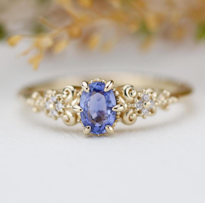 An elegant vintage-inspired 18k yellow gold ring featuring an oval natural blue sapphire as the center piece, accented by sparkling natural diamonds in a delicate floral and scroll design.